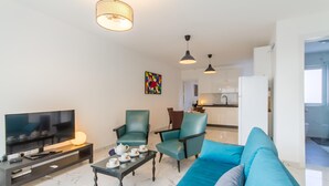 Ein modernes Wohnzimmer mit Flachbildfernseher, einer Couch, zwei Sesseln, einem Couchtisch und einer Küche im Hintergrund.