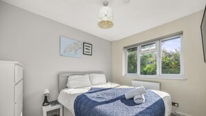 A bedroom with a bed, a nightstand, a lamp, a map, and a window with a view of greenery. - Secret Garden House With Free Parking By My Getaways (Horsham)