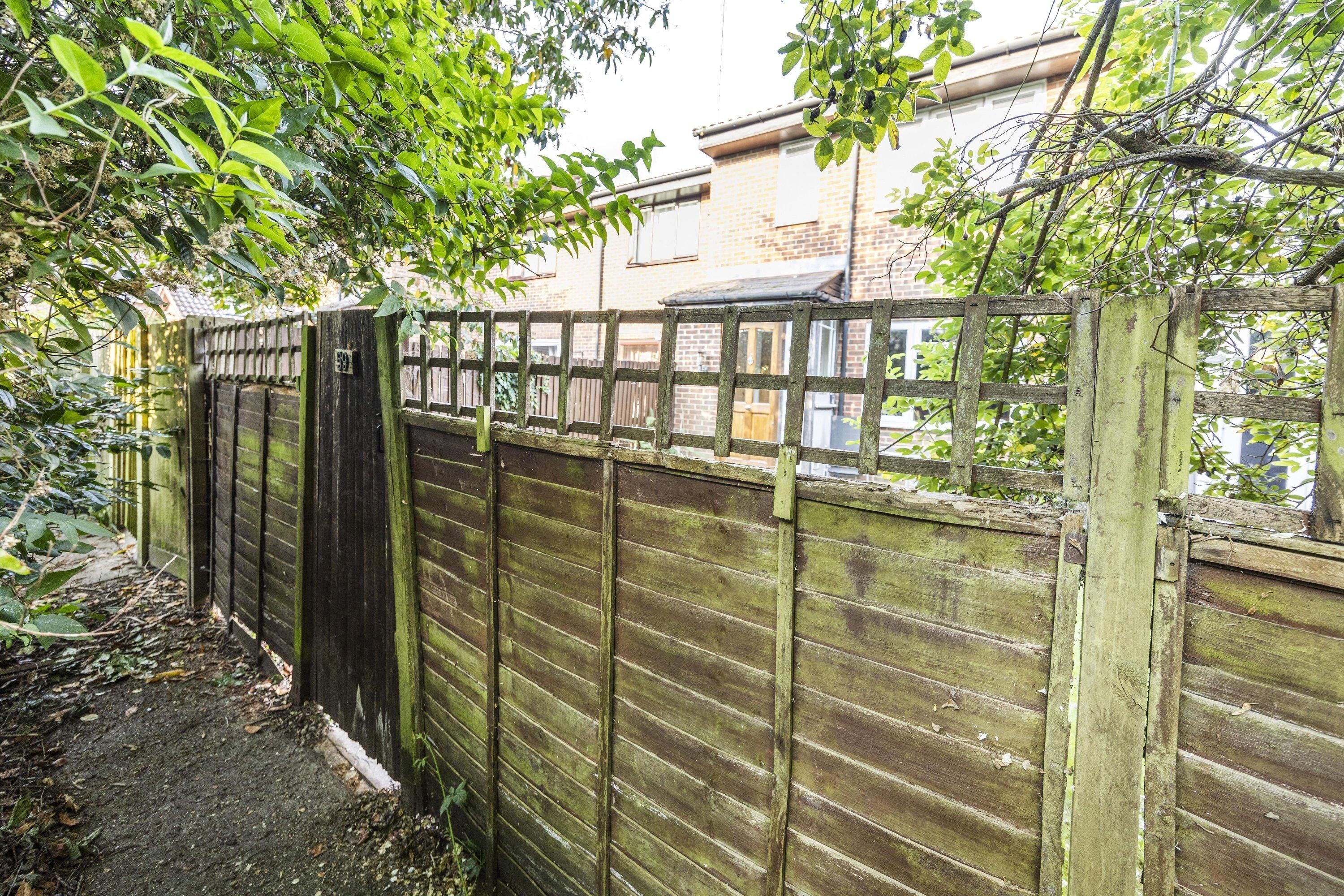 A wooden fence with a gate, surrounded by greenery and a building in the background.