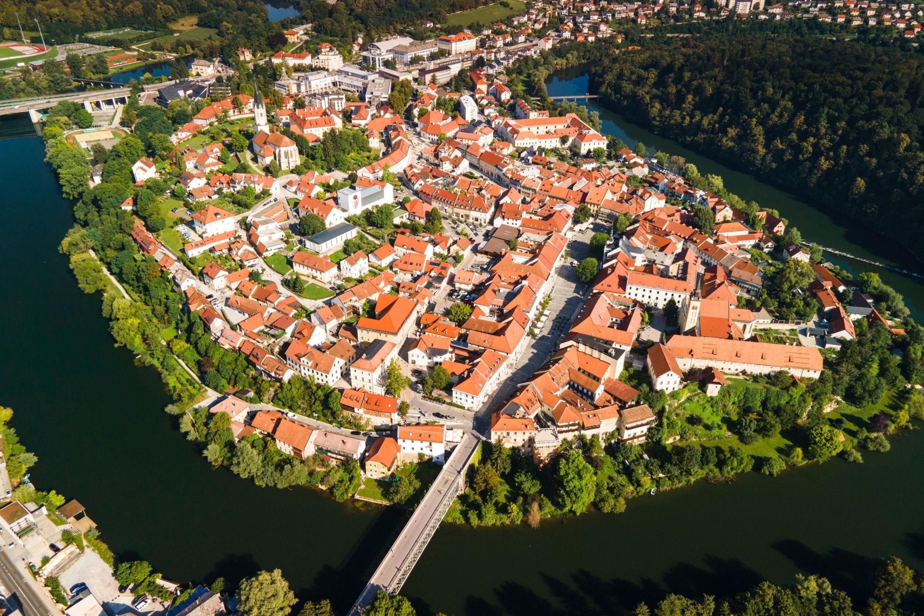 An aerial view of a historic town with a river, featuring a central square, red-roofed buildings, and a bridge.