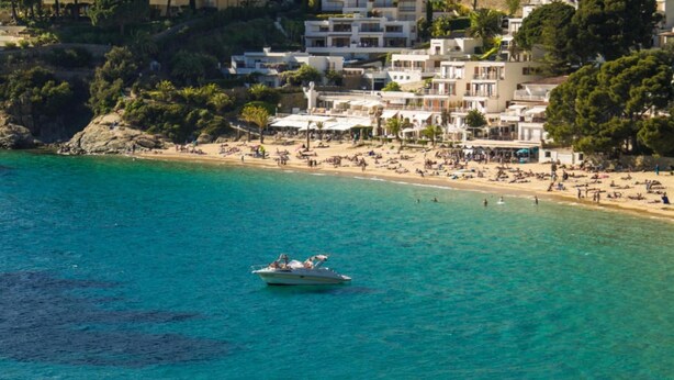 A coastal area with a boat on the water, a beach with people, and buildings in the background. - Arago 273 in Roses With 2 Bedrooms and 2 Bathrooms (Roses)