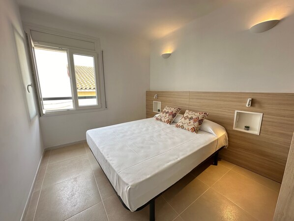 A bedroom with a bed, two pillows, a nightstand, a wall-mounted light, and a window.
