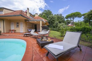 A poolside area with lounge chairs, a table with drinks, and a house in the background. - Villa Piero With Pool (Pietrasanta)