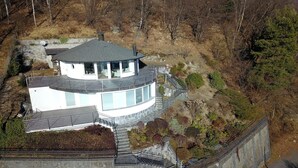Uma casa moderna e circular com uma grande fachada de vidro, rodeada por um muro de pedra e uma encosta arborizada.
