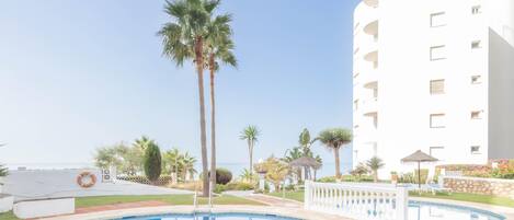 Une piscine entourée d’une clôture blanche, avec des palmiers et un bâtiment blanc doté de balcons.