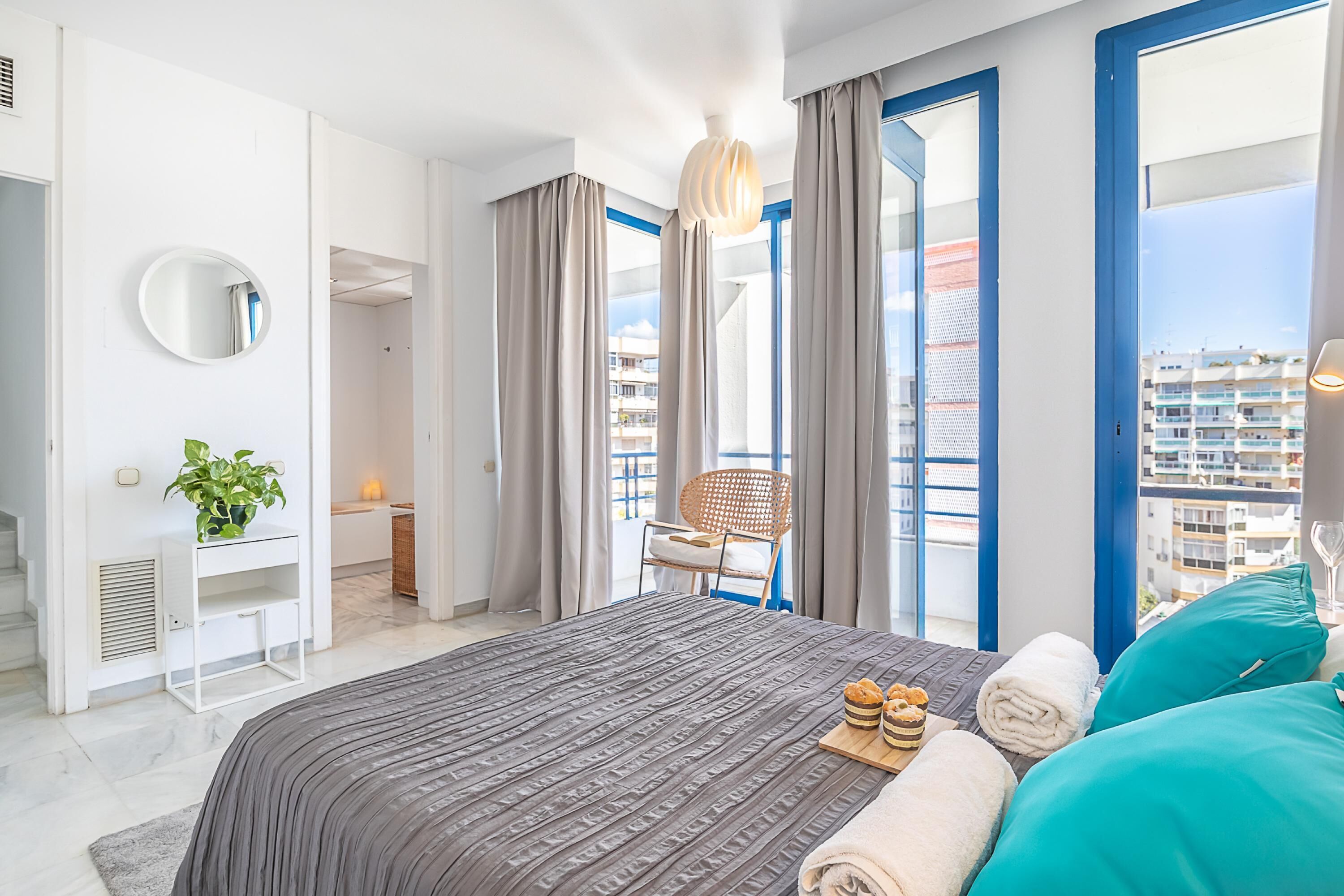 A modern hotel room with a large bed, a desk, a chair, and a view of the city through large windows.