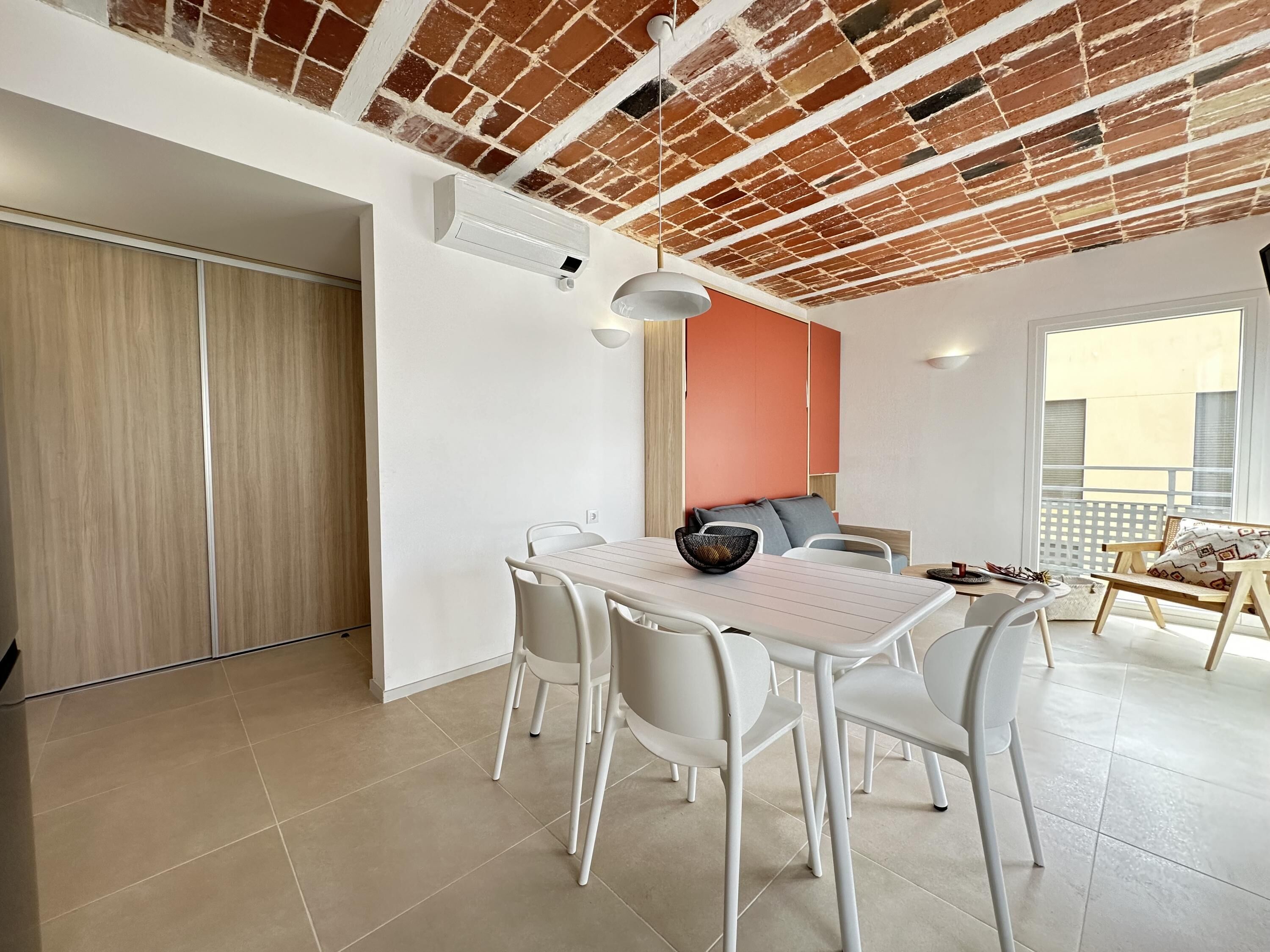 A dining area with white chairs and a table, a brick ceiling, and a view of the outside balcony.
