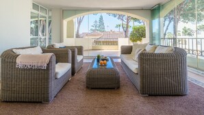 A spacious living area with wicker furniture, a coffee table with fruits, and a view of a balcony with trees and a building in the background. - Hacienda Playa Beach And Pool (Marbella)