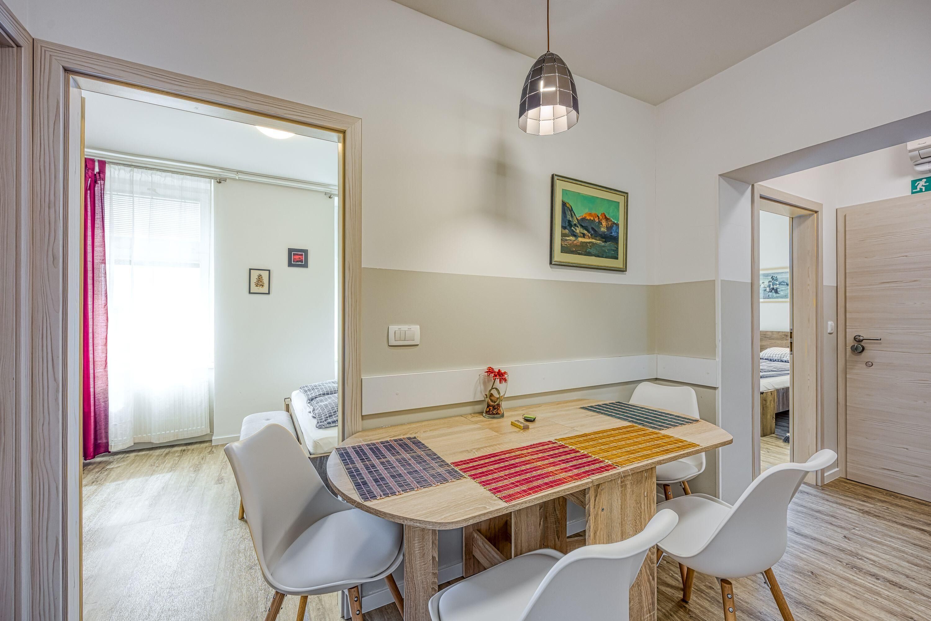 Un comedor con una mesa de madera, sillas blancas y manteles individuales de colores. Hay una lámpara colgante sobre la mesa y un cuadro en la pared.