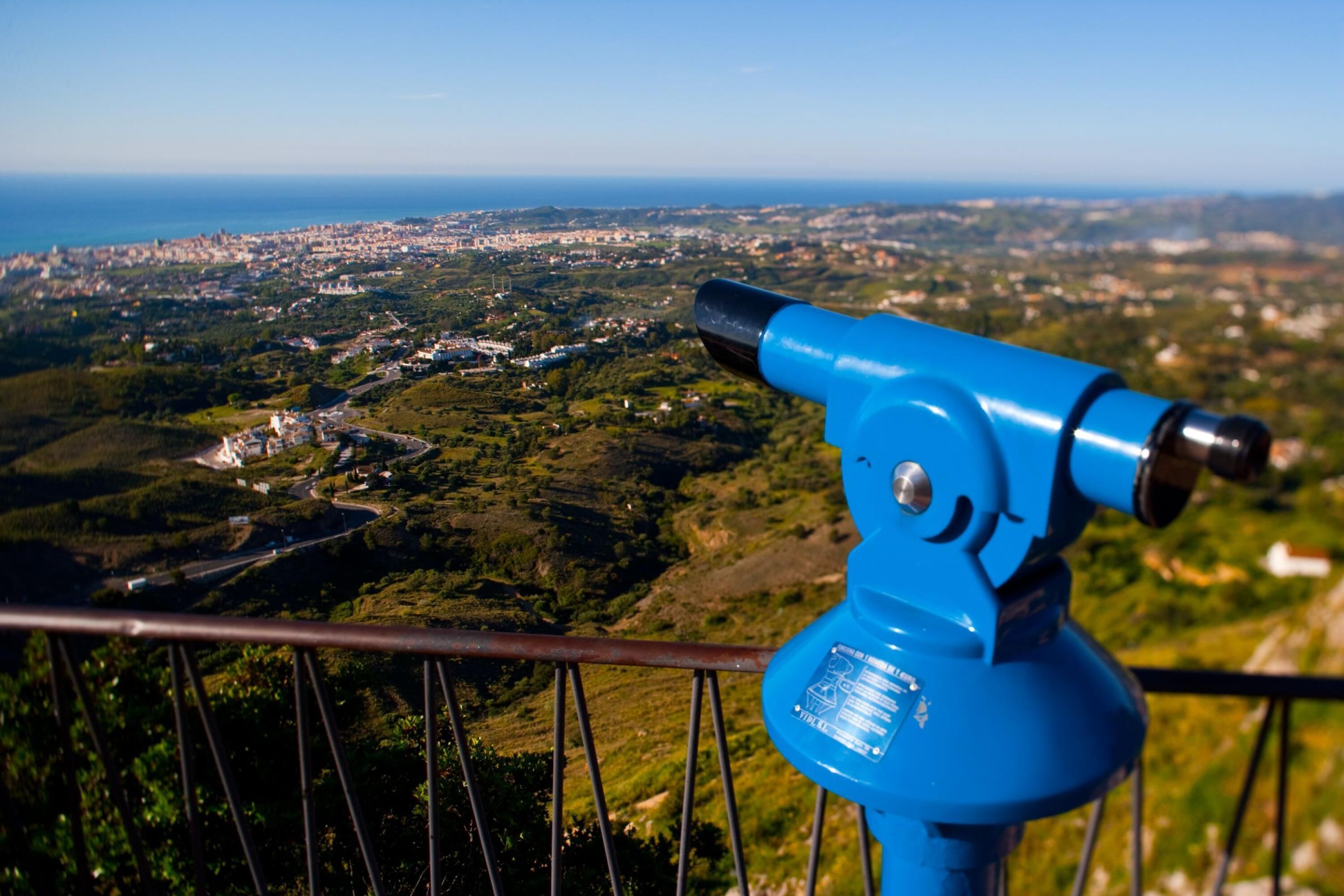 Ein Teleskop auf einer Geländer mit Blick auf eine Küstenlandschaft, einschließlich Gebäude und Grünflächen.