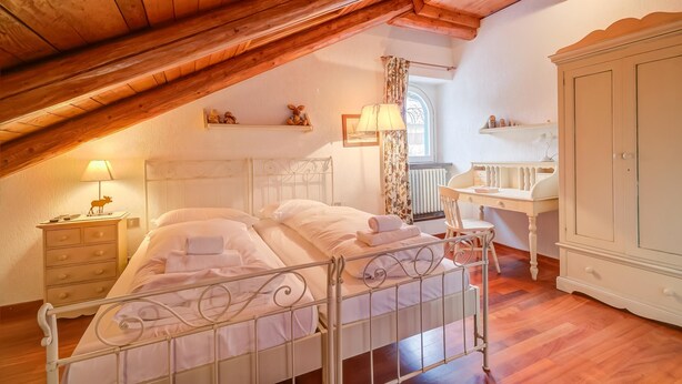 Una camera da letto mansardata accogliente, con travi a vista, un letto con lenzuola bianche, un comodino in legno, una scrivania e un armadio.