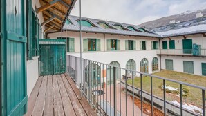 Un edificio con un balcone in legno, persiane verdi e vista su un cortile con una montagna innevata sullo sfondo.