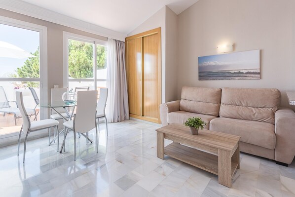 A modern living room with a beige sofa, a glass coffee table, and a dining area with white chairs and a glass table. - Marbella Resort 8 Pool Whirlpool Sauna (Marbella)