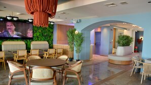 Un moderno vestíbulo de hotel con una mesa redonda de madera, sillas de mimbre, un televisor de pantalla plana grande, plantas verdes y un área de descanso con un sofá blanco y una planta en maceta.