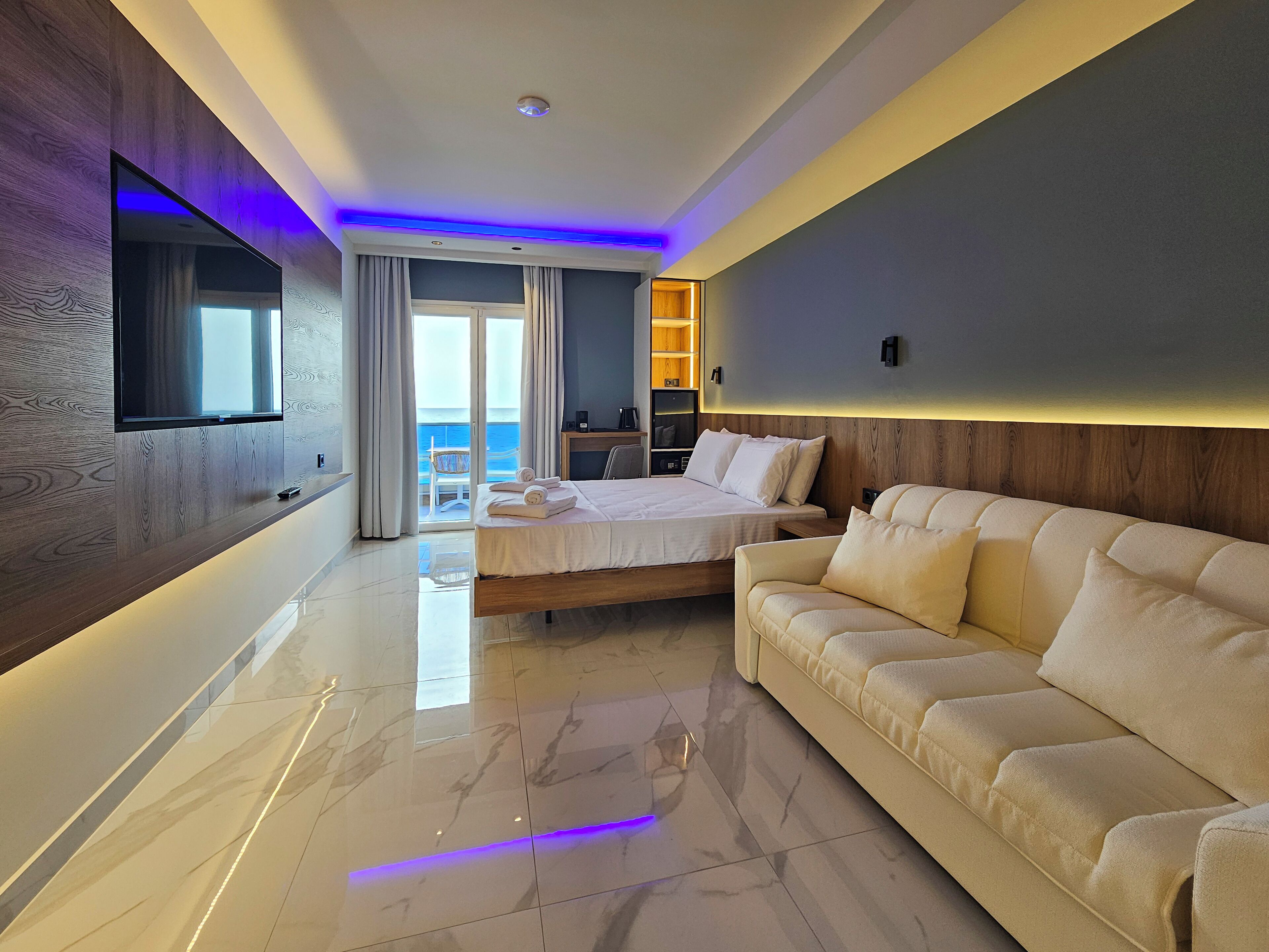 Quarto de hotel moderno com uma cama grande, um sofá, uma televisão fixada na parede e vista para o mar através da porta de correr de vidro.