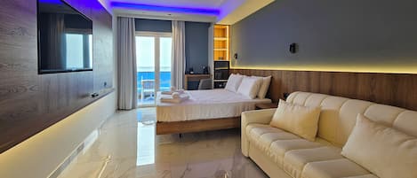 A modern hotel room with a large bed, a sofa, a TV mounted on the wall, and a view of the sea through the sliding glass door.