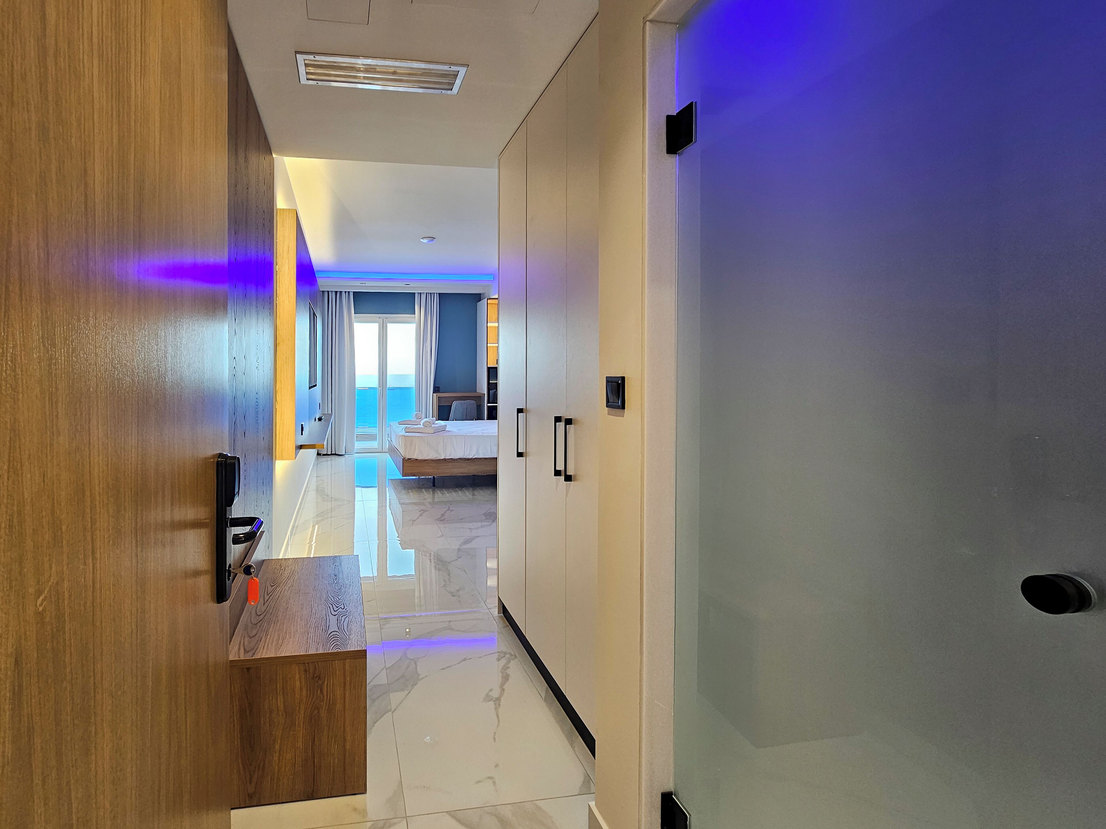 A modern hotel room with a wooden desk, a bed with white linens, a bathroom with a shower, and a view of the sea.