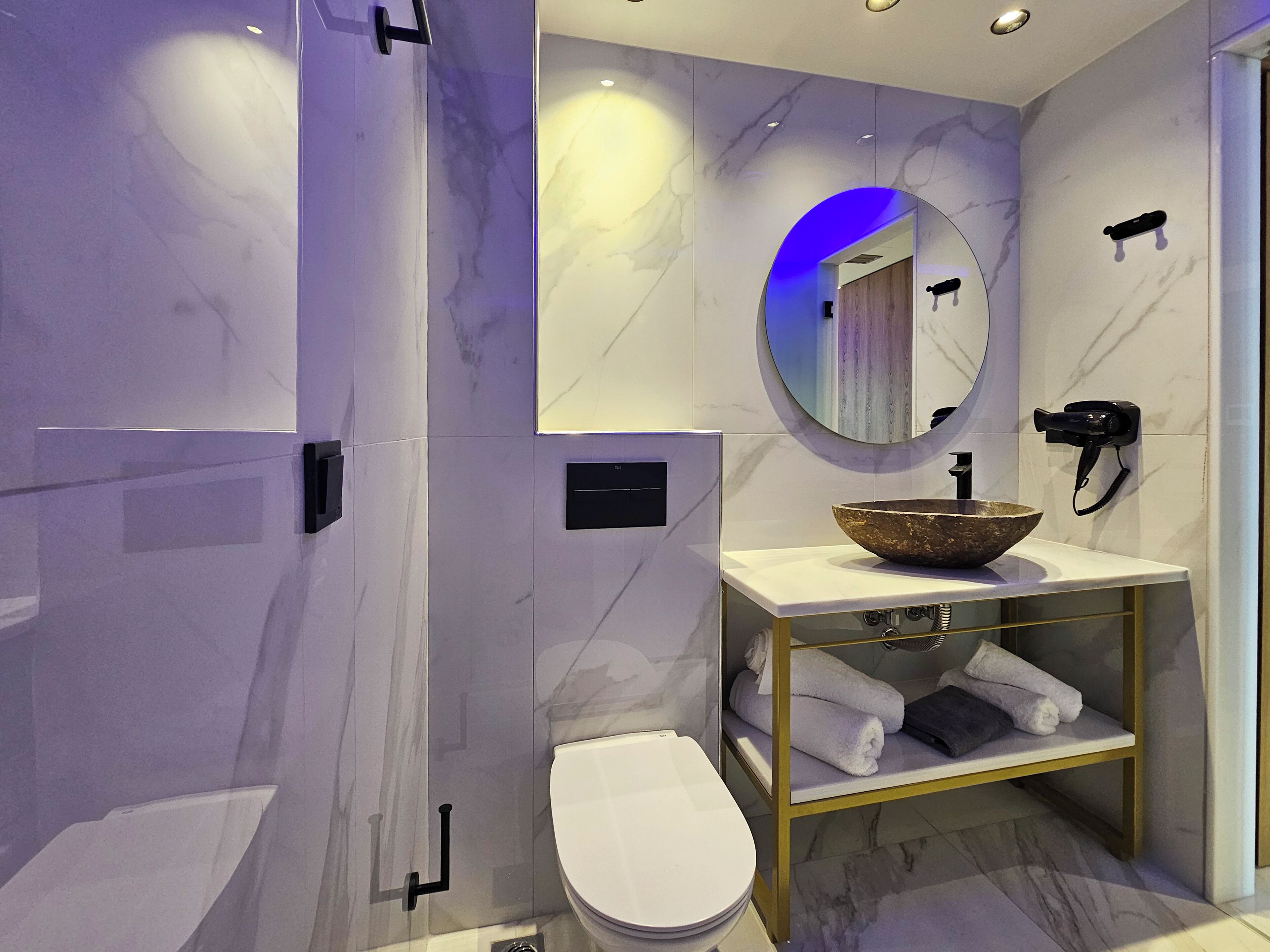 Un bagno moderno con parete in marmo, un WC bianco, un mobile lavabo con lavandino capiente e uno specchio con illuminazione blu, oltre a un porta asciugamani.