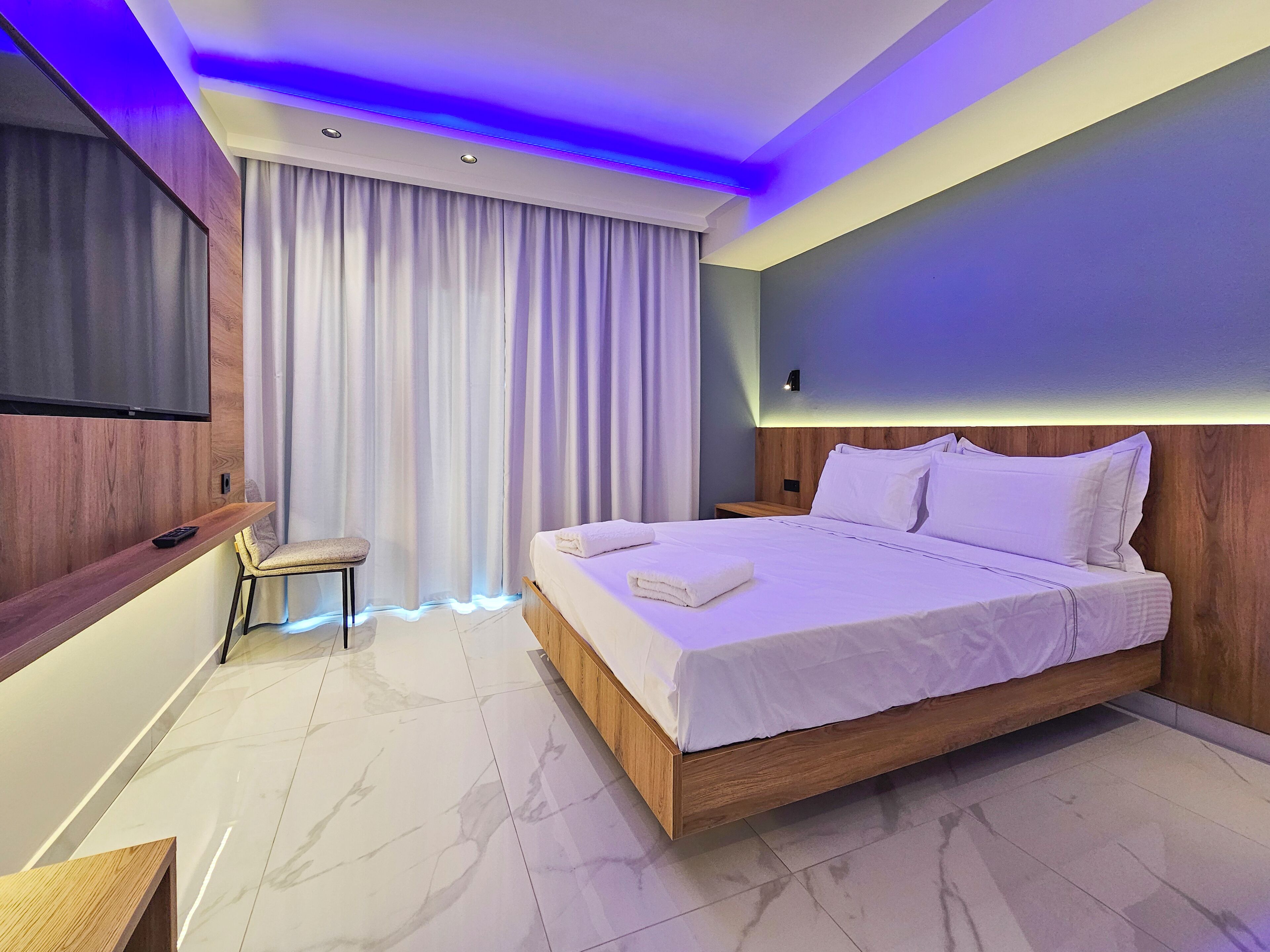 A modern hotel room with a large bed, a TV mounted on the wall, a small seating area, and a large window with curtains.