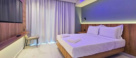 Una habitación de hotel moderna con una cama grande, un televisor montado en la pared, un pequeño rincón de descanso y un amplio ventanal con cortinas.