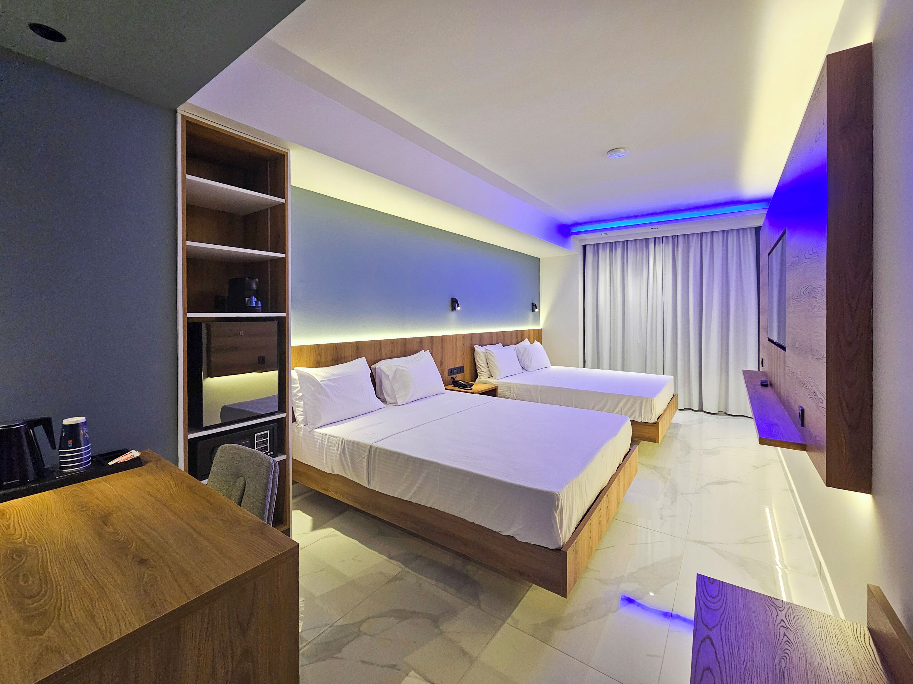 A hotel room with two beds, a wooden desk, a built-in shelf, and a large window with curtains.