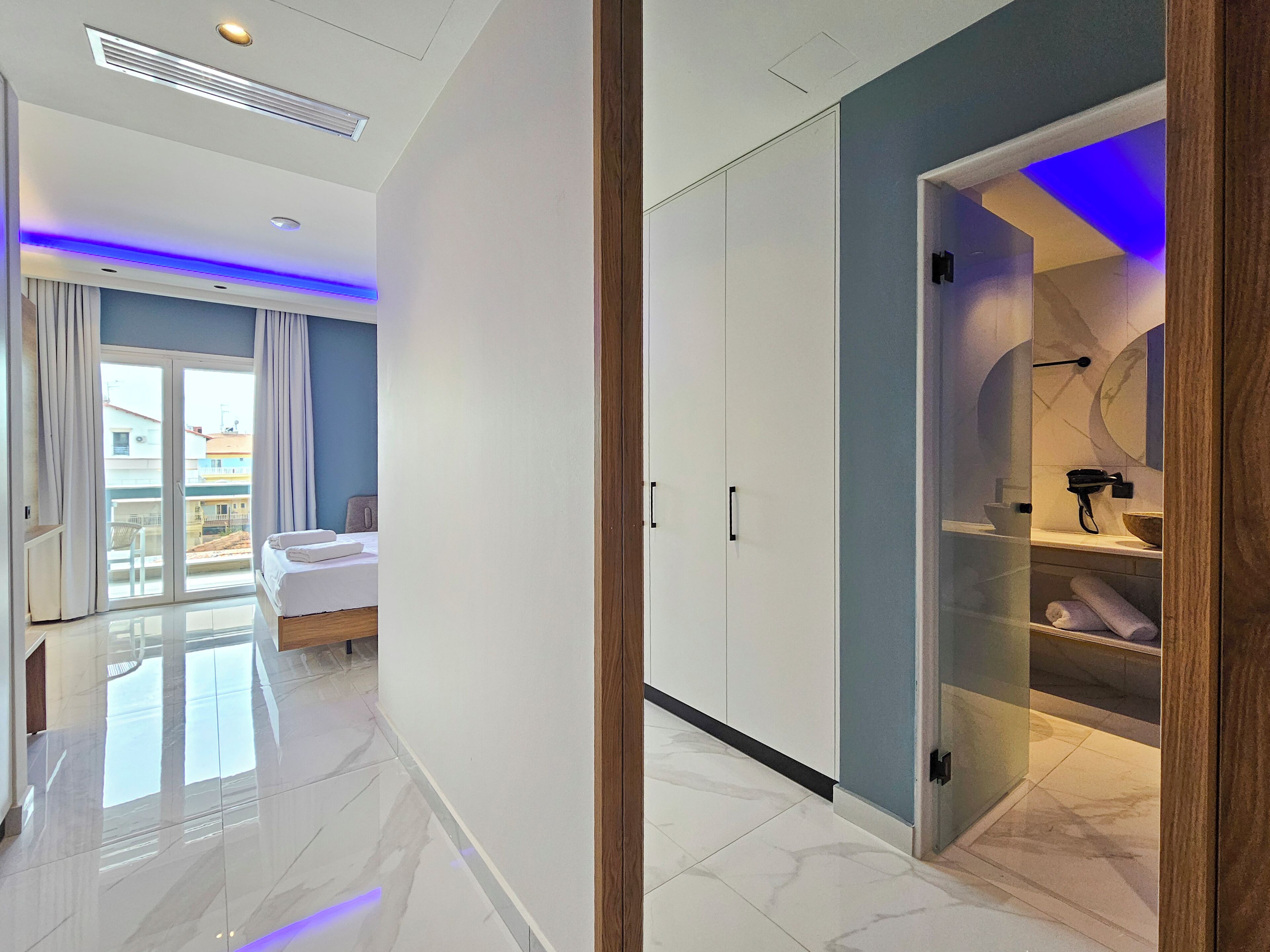 A modern hotel room with a large bed, a sliding glass door to a balcony, a walk-in closet, and a bathroom with a vanity and towel rack.