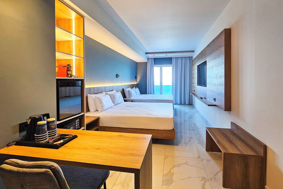Habitación de hotel con dos camas, un escritorio con cafetera, televisor y vista al mar.