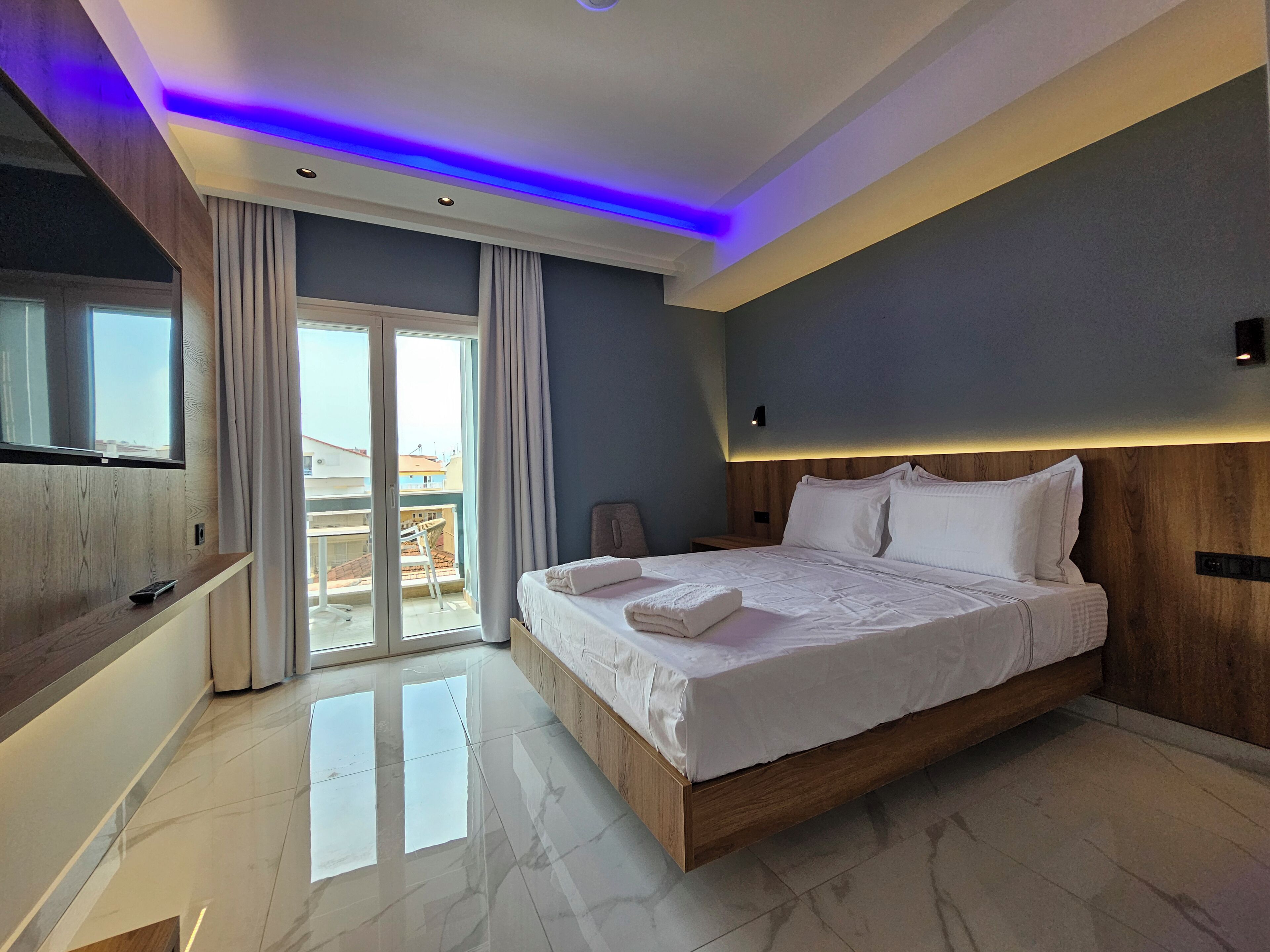 A modern bedroom with a large bed, a TV mounted on a wooden panel, and a balcony with curtains.