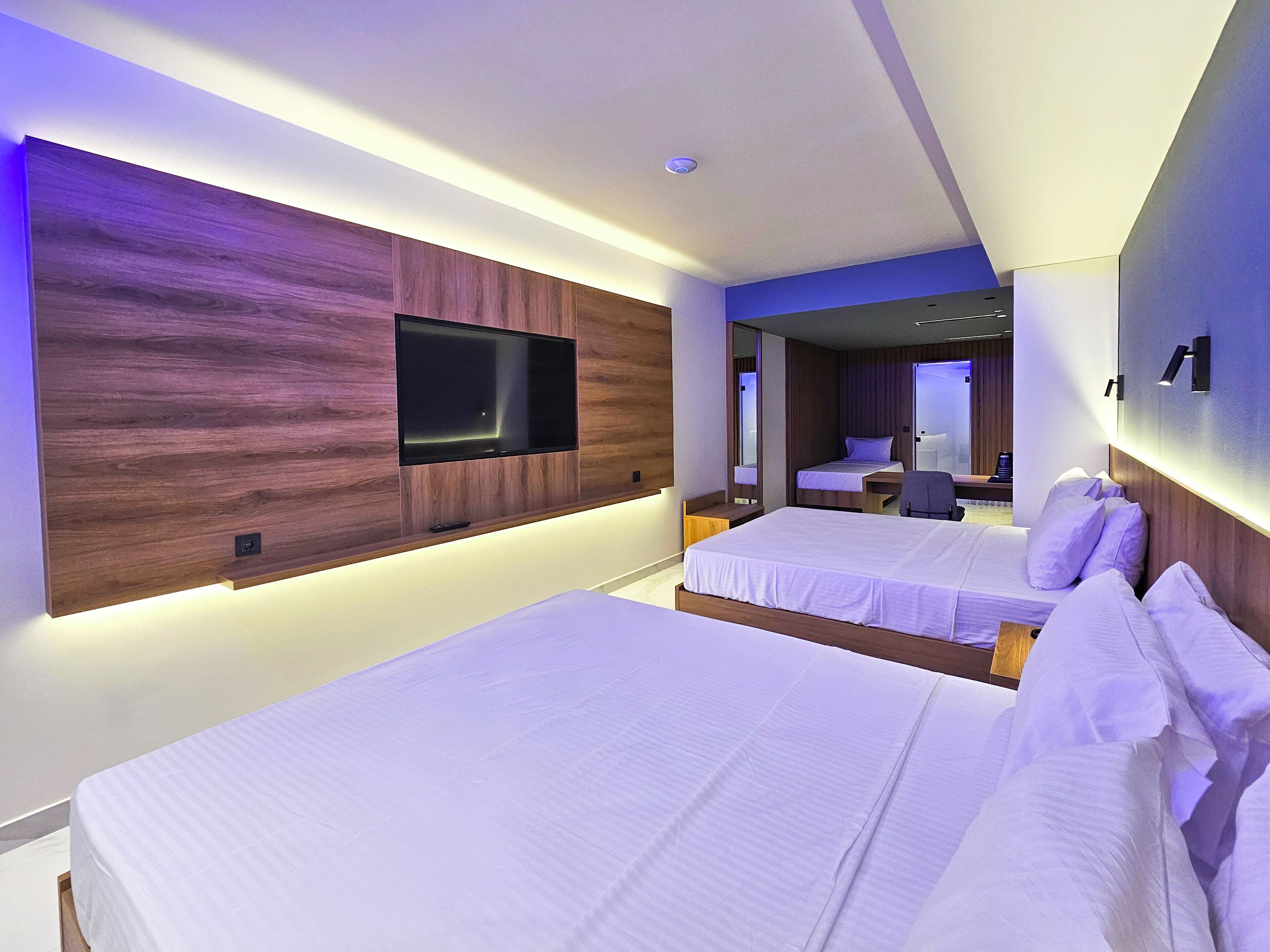 Habitación de hotel moderna con dos camas, un televisor de pantalla plana montado en un panel de madera y un espejo sobre la cama.