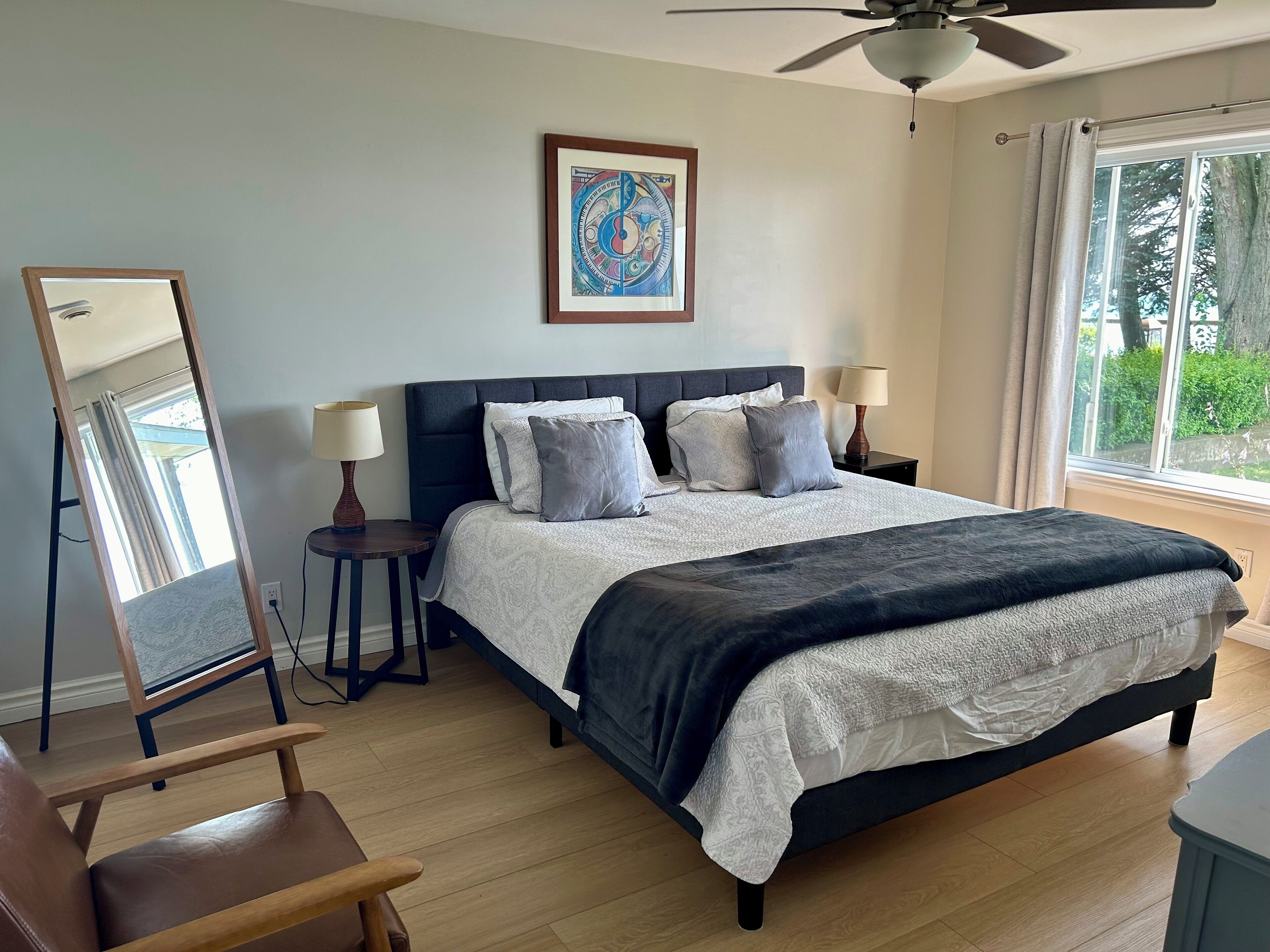 A bedroom with a bed, two bedside tables with lamps, a large mirror, a ceiling fan, and a window with curtains.