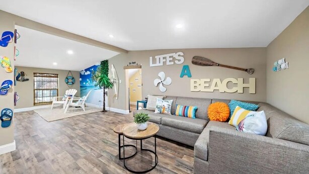 Un salón con sofá gris, una mesita de centro y una pared decorada con el texto 'LIFE'S A BEACH'.