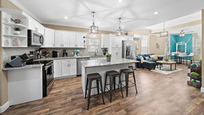 Una cocina moderna con armarios blancos, una isla grande con taburetes de bar y una zona de comedor con una pared de acento color aguamarina.