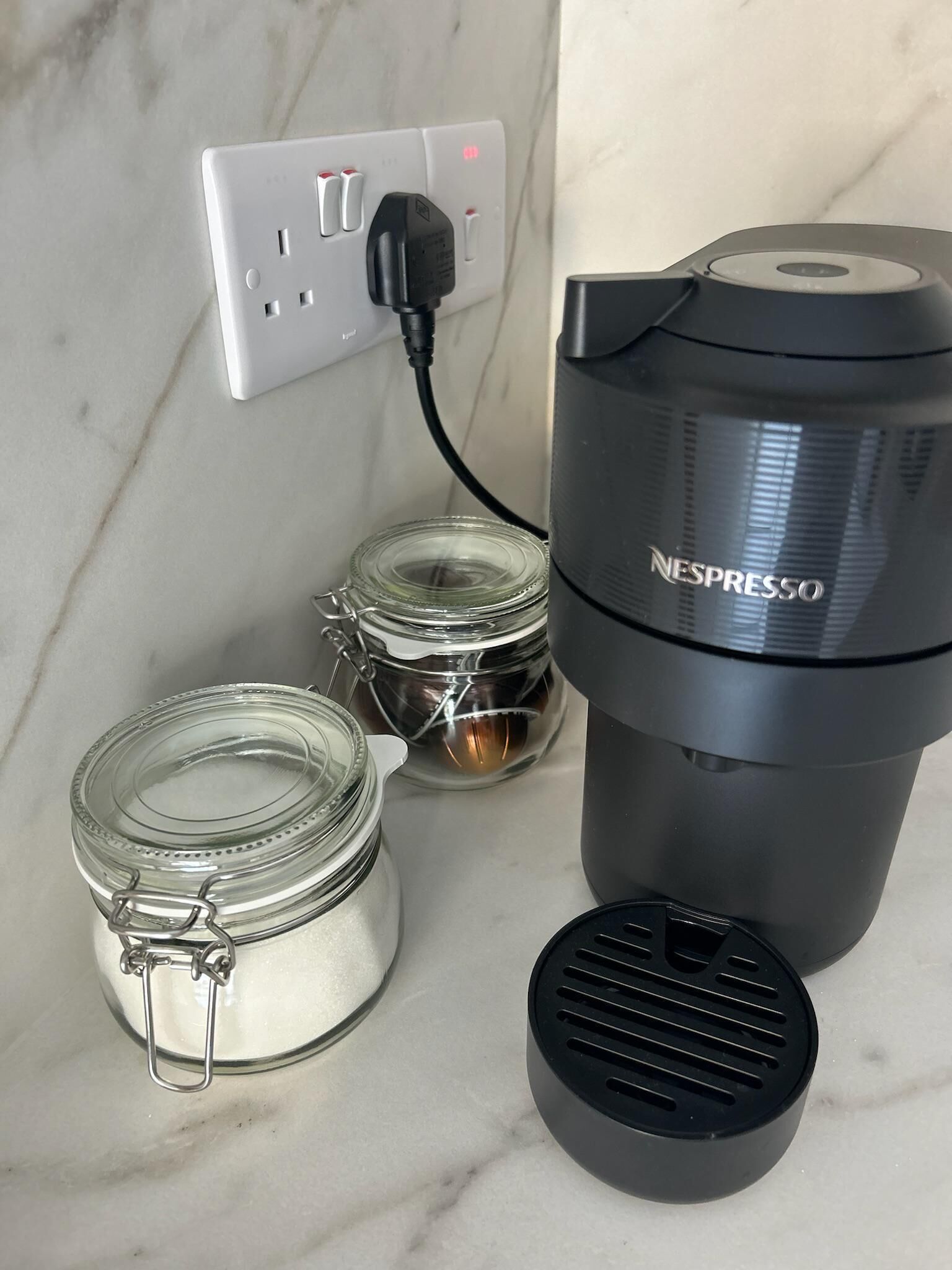 Une machine à café Nespresso, un bocal en verre avec un fermoir en métal et un récipient noir doté d’une ouverture d’aération, le tout posé sur un comptoir en marbre.
