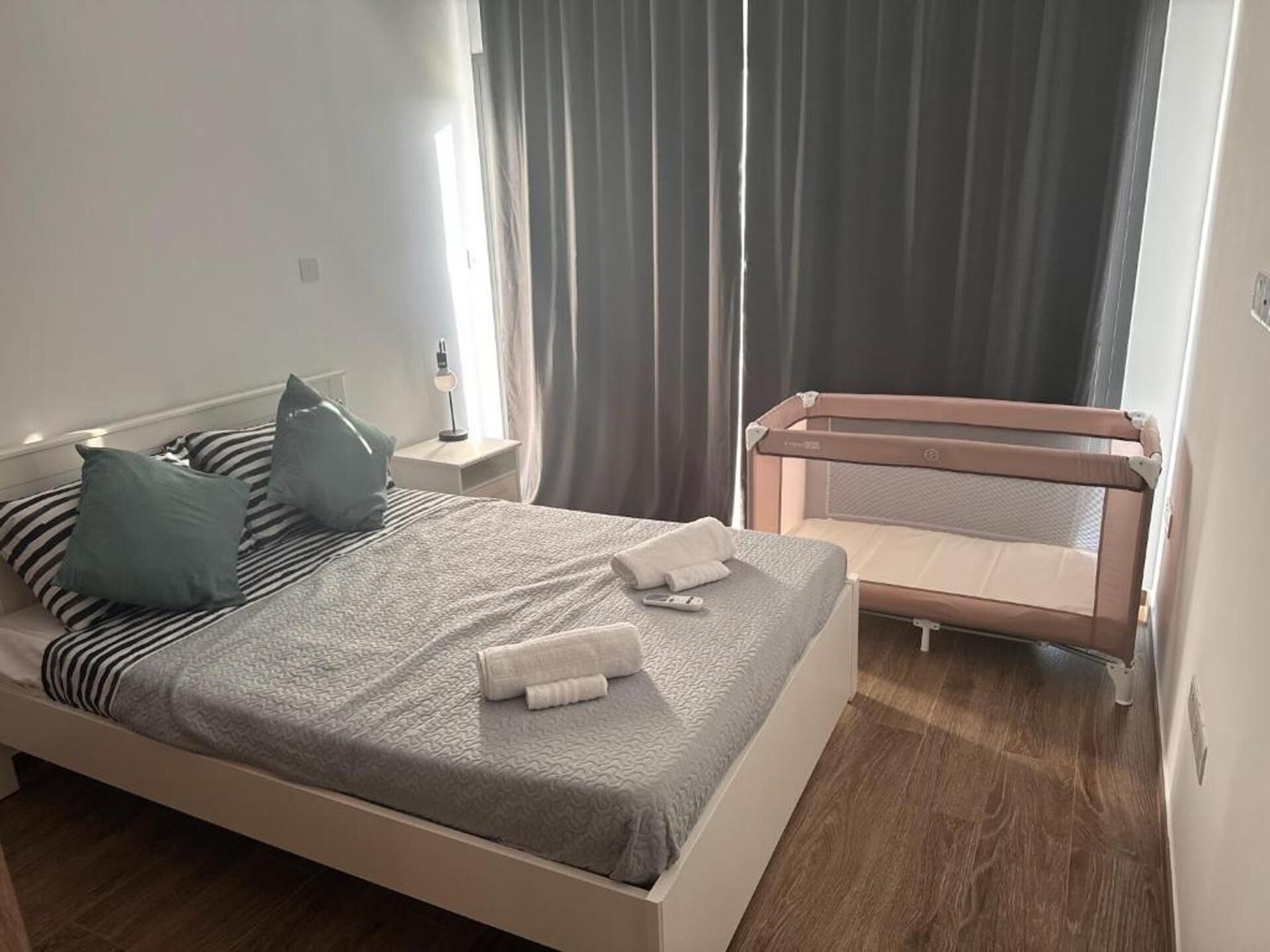 Una cama bien tendida con ropa de cama a rayas y almohadas, una mesita de noche con lámpara, y un pequeño balcón con barandilla.