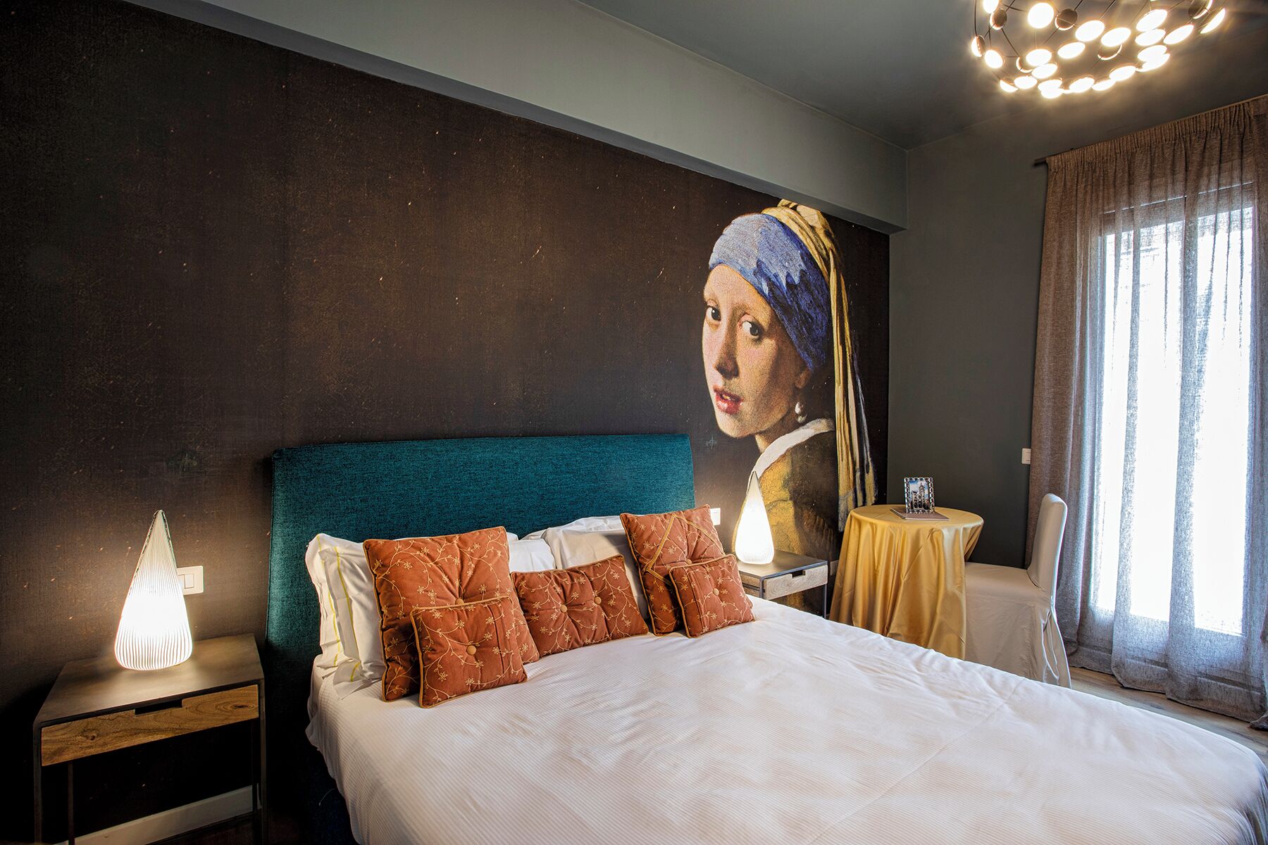 A bedroom with a large bed, a nightstand with a lamp, a table with a picture frame, and a window with curtains.