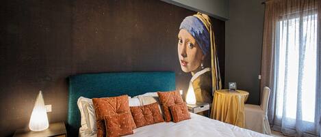 Un dormitorio con una cama grande, una mesita de noche con una lámpara, una mesa con un marco de fotos y una ventana con cortinas.