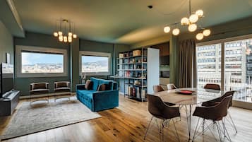 A modern living room with a blue sofa, wooden flooring, a dining area with a table and chairs, a bookshelf, and large windows offering city views.