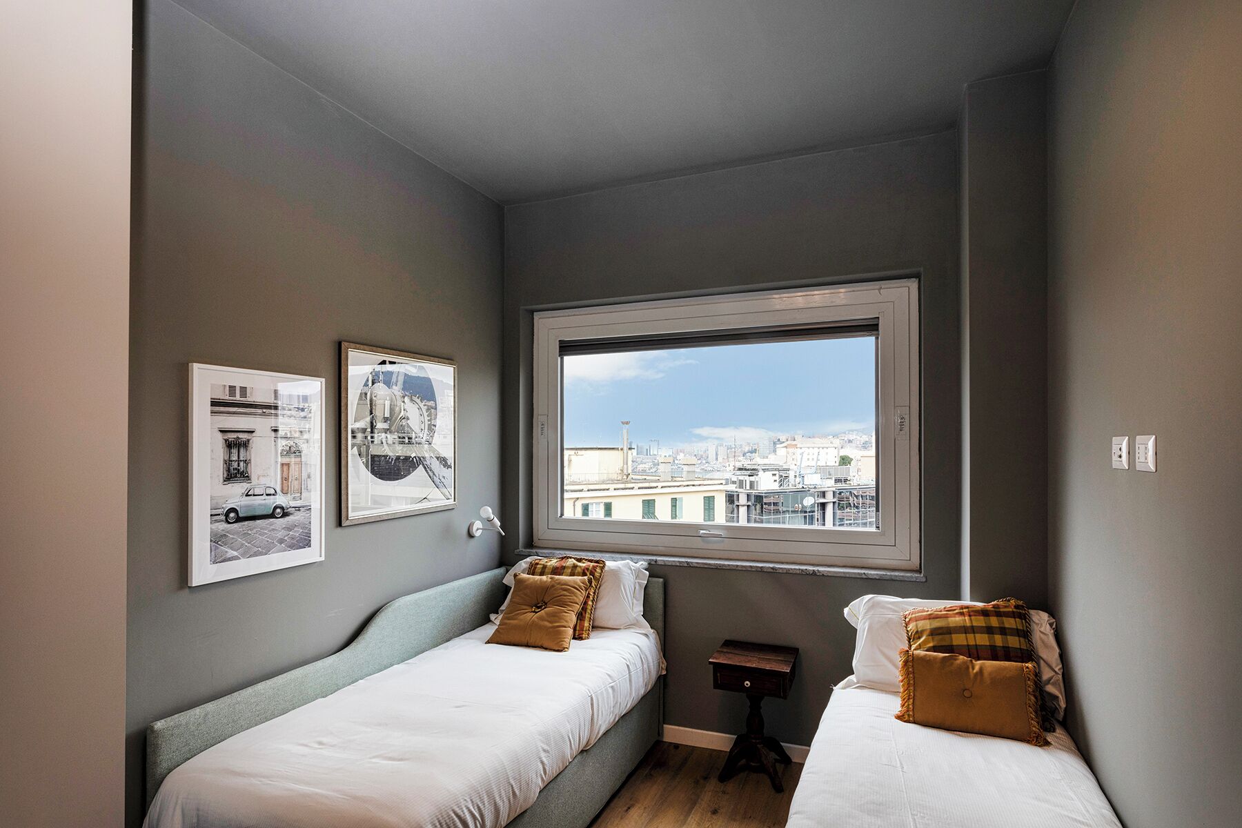 Quarto compacto com duas camas de solteiro, uma mesinha de cabeceira e janela com vista para a cidade.