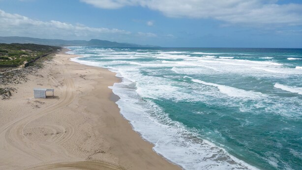 A beach with a sandy shore, a small structure, and waves crashing onto the beach. - Nicole House With Stunning View And Pool (Badesi)