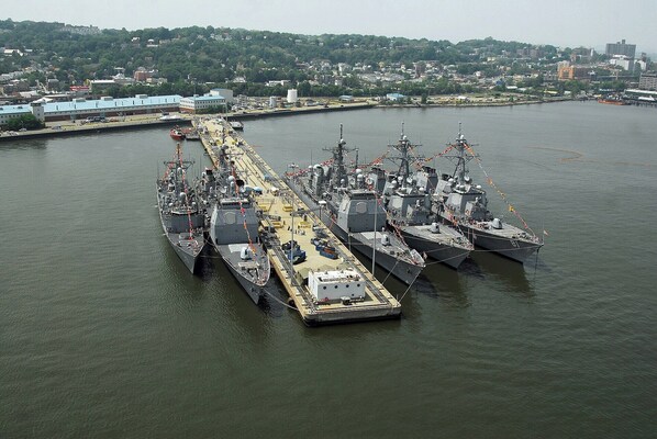 A large naval ship docked at a port with a cityscape in the background.