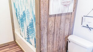 A bathroom with a wooden floor, a white toilet, a shower curtain with a forest design, and a wall-mounted towel rack.