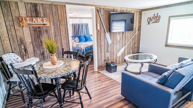A room with a wooden wall, a round table with chairs, a sofa, a TV, and a sign that says 'LAKE'.
