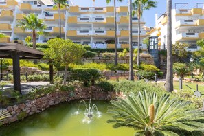 Surrounded by lush greenery and palm trees, this tranquil resort area features a serene pond and a modern building with spacious balconies.