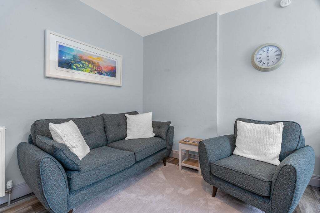 Ett vardagsrum med en grå soffa, en mindre grå fåtölj, en inramad tavla och en väggklocka.