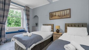 A hotel room with two beds, a window with curtains, a painting on the wall, and a small bedside table with a lamp.