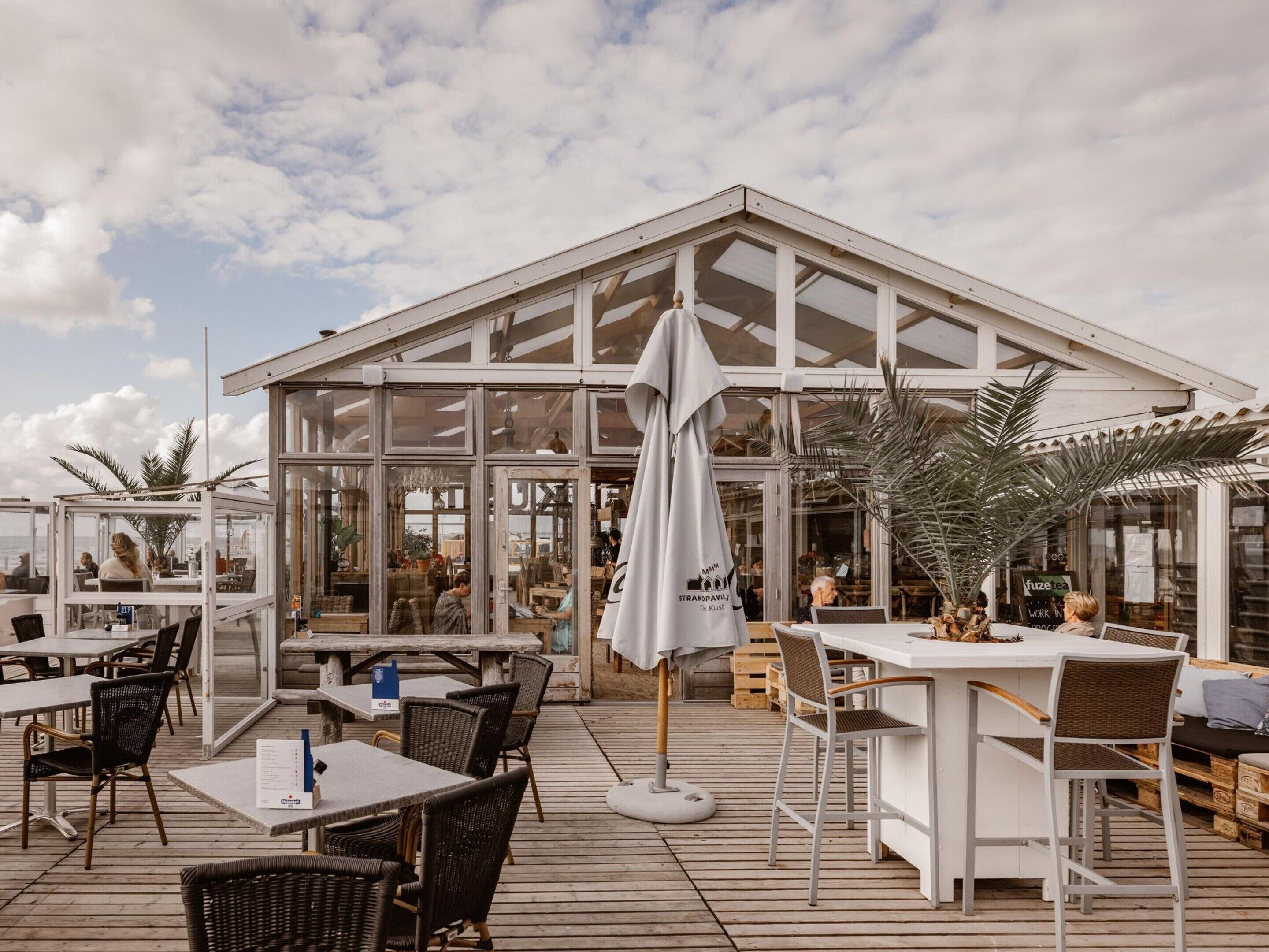 En udendørs café med borde og stole, en stor parasol og udsigt til havet.