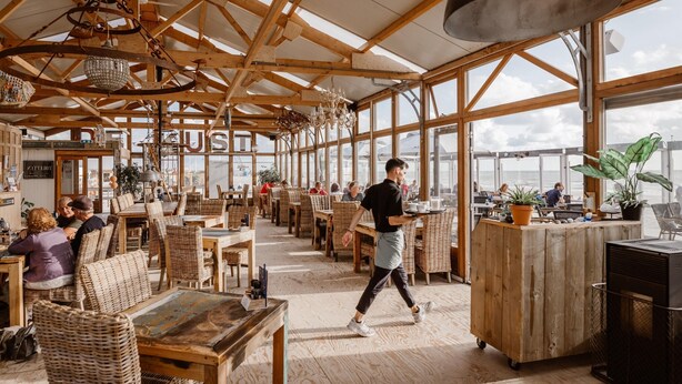Ein geräumiges Café mit Holzbalken, Korbmöbeln und großen Fenstern, die den Blick auf den Himmel ermöglichen.