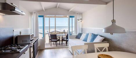 Eine moderne Küche mit Gasherd, weißen Schränken und einem Essbereich mit rundem Tisch und Stühlen. Durch eine Glastür bietet sich Blick auf den Strand.