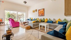 Un salón amplio con un sofá grande, un sillón rosa y una mesa de centro de madera. Una ventana grande ofrece vistas al exterior.