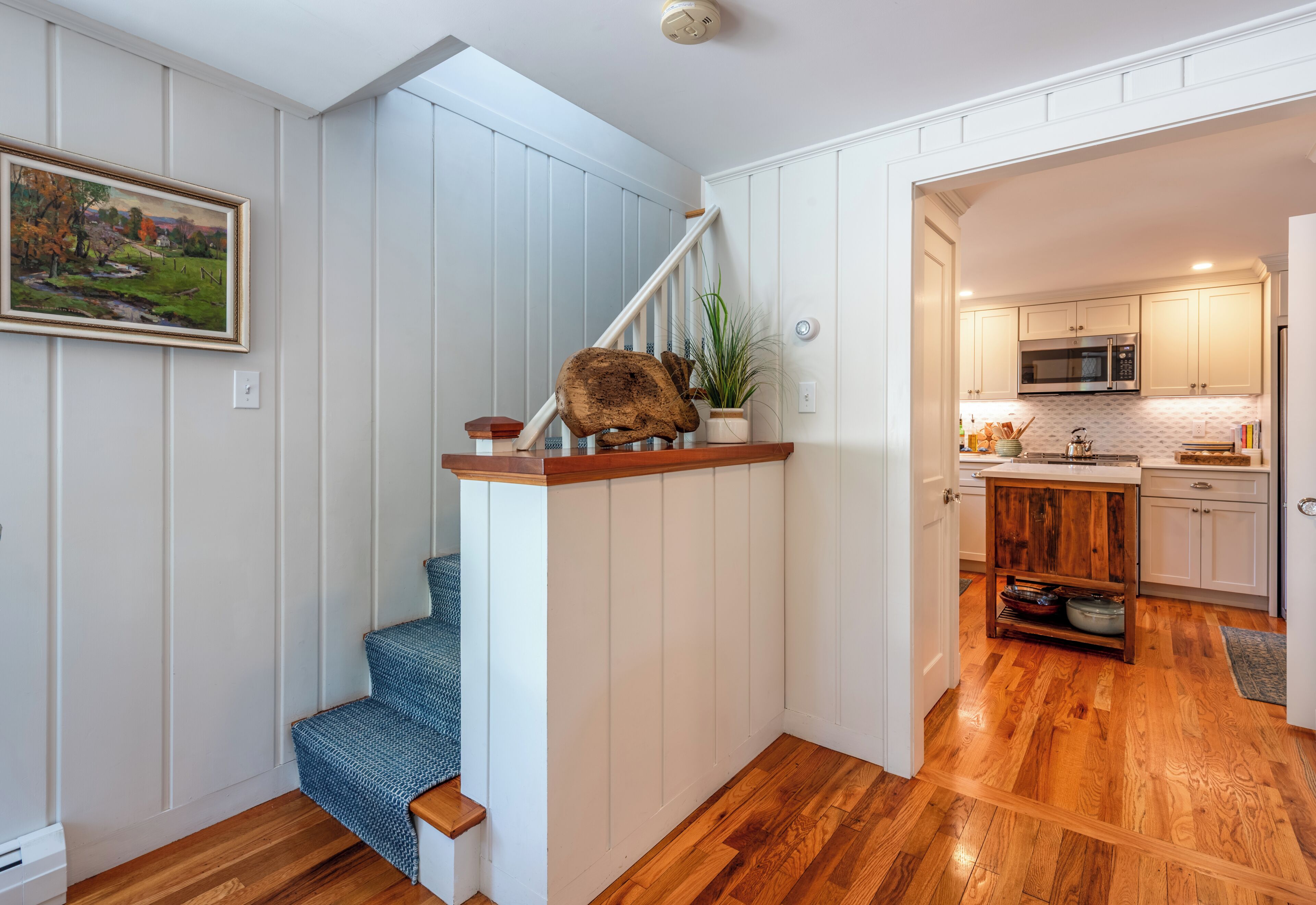 A staircase with a wooden handrail and a blue patterned chair at the bottom. A kitchen with white cabinets and a microwave is visible in the background.
